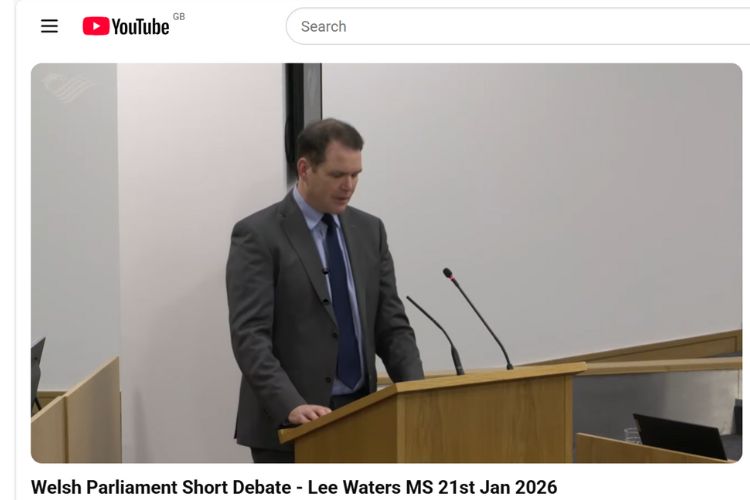 A screenshot of an online video featuring a man standing at a microphone addressing a room full of people